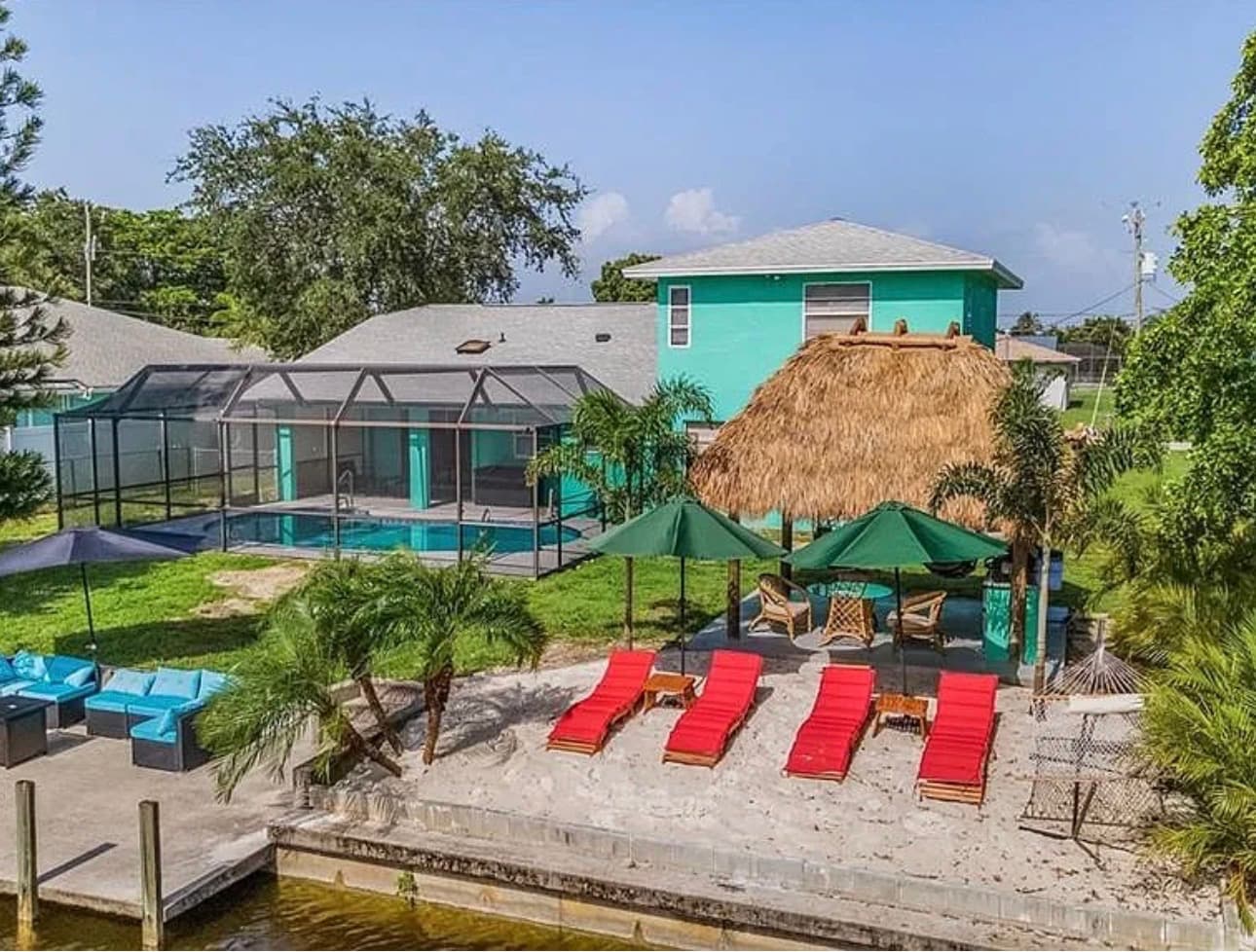 Canal-front vacation home with screened pool, tiki hut, sand beach area, and red loungers